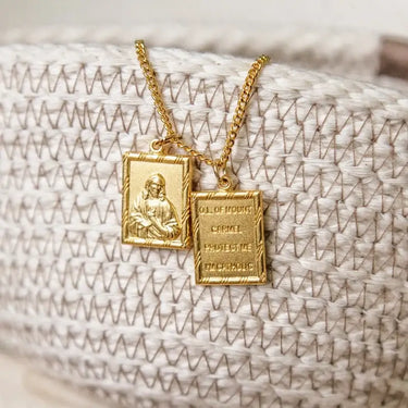 Two gold-toned religious pendant necklaces displayed on a woven cream fabric surface. One rectangular pendant shows an embossed figure, and the other rectangular pendant has engraved text including 'OJ.