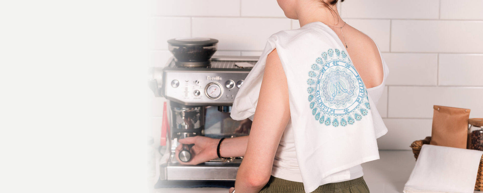 Person using a coffee machine in a kitchen setting with a dish towel draped over their shoulder.