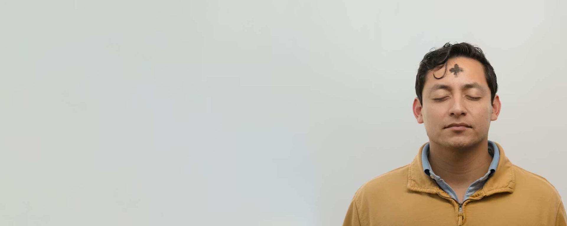 Man with eyes closed and an ash cross on his forehead against a plain background.