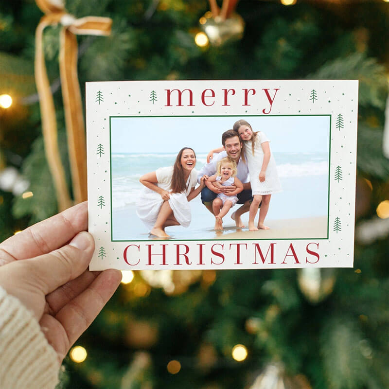 Hand holding a Christmas card with a family photo against a Christmas tree background.