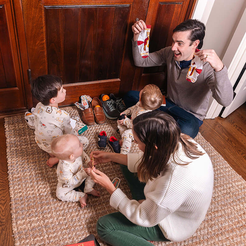 Family of four with three young children on a carpeted floor with father showing off Christmas socks.