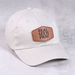 White cap with a brown leather patch featuring 'Real Men Pray' on a light gray background.