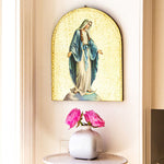 Decorative shelf with a religious icon of a woman in a blue robe and white dress, a white vase with pink flowers, and a light-colored wall.