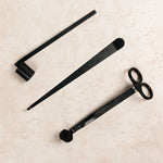 A black candle snuffer, candle pick and candle scissors on a beige surface.