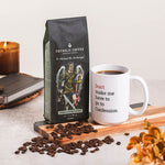 Catholic Coffee bag with a St. Michael the Archangel design next to a white mug with text, on a table with coffee beans and a candle.