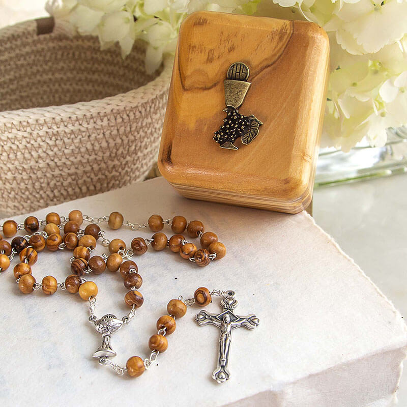 Wooden rosary with silver crucifix on a marble surface with a wooden box and flowers in the background.