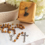 Wooden rosary with silver crucifix on a marble surface with a wooden box and flowers in the background.