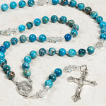 Blue beaded rosary with silver crucifix and Saint pendant on a light background.