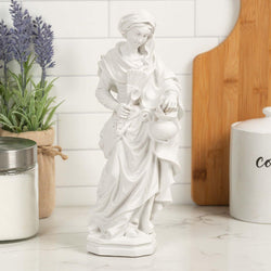White statue of a woman holding a jug, a spoon and a sweeper on a marble surface with a plant and cutting board in the background.