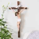 Wooden cross with a figure of Jesus on a white wall, next to a plant.