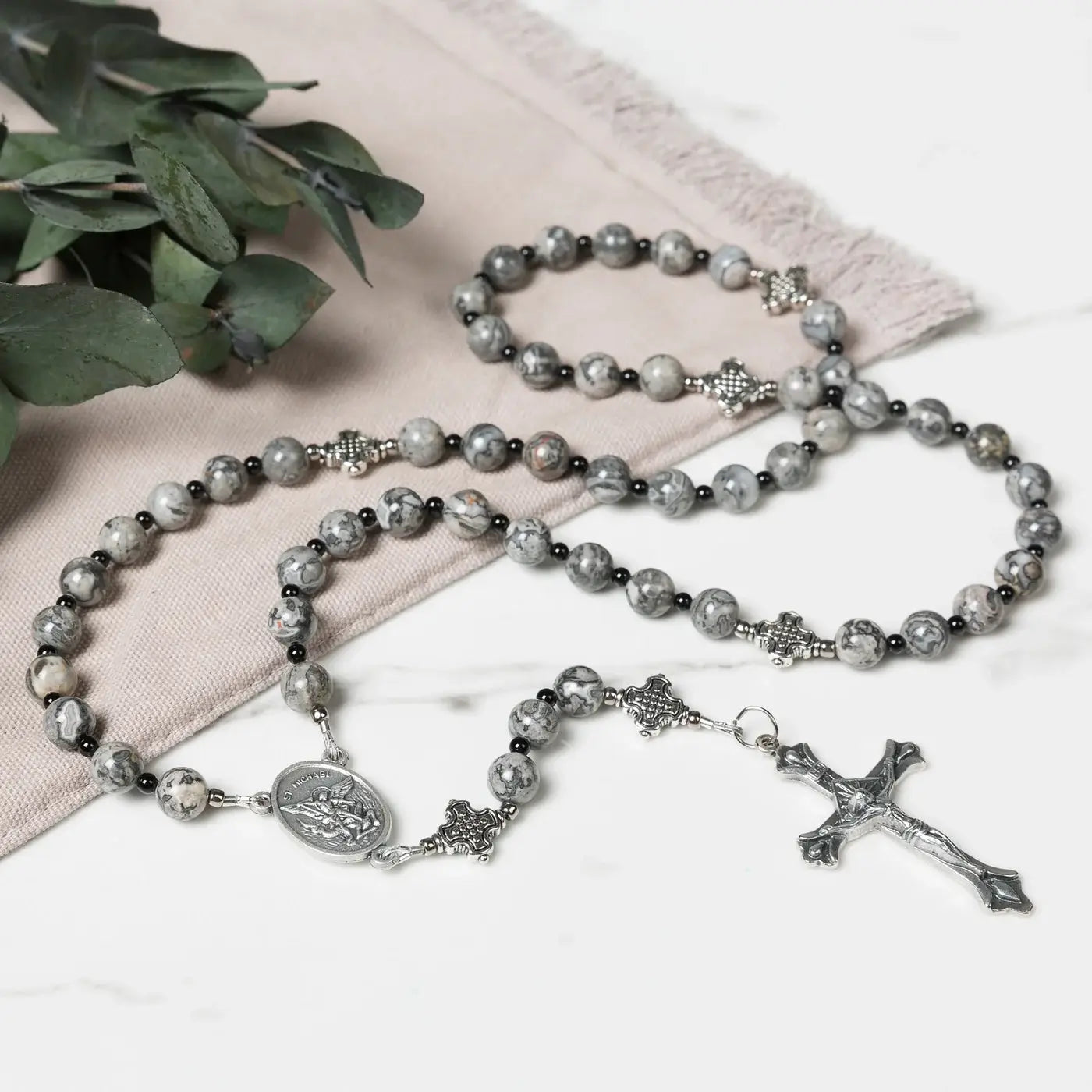 Rosary with gray beads and silver cross on a white surface with green leaves.
