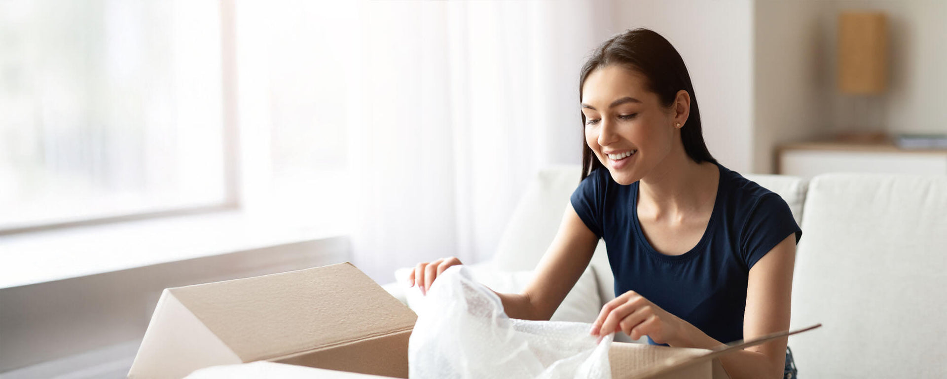 Smiling woman sitting on a couch opening a box in a bright room.