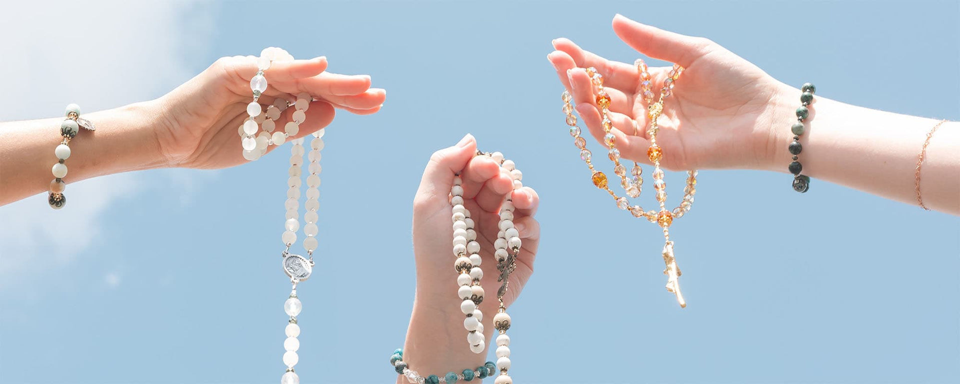 Three hands holding white and gold beaded rosaries against a clear blue sky background.