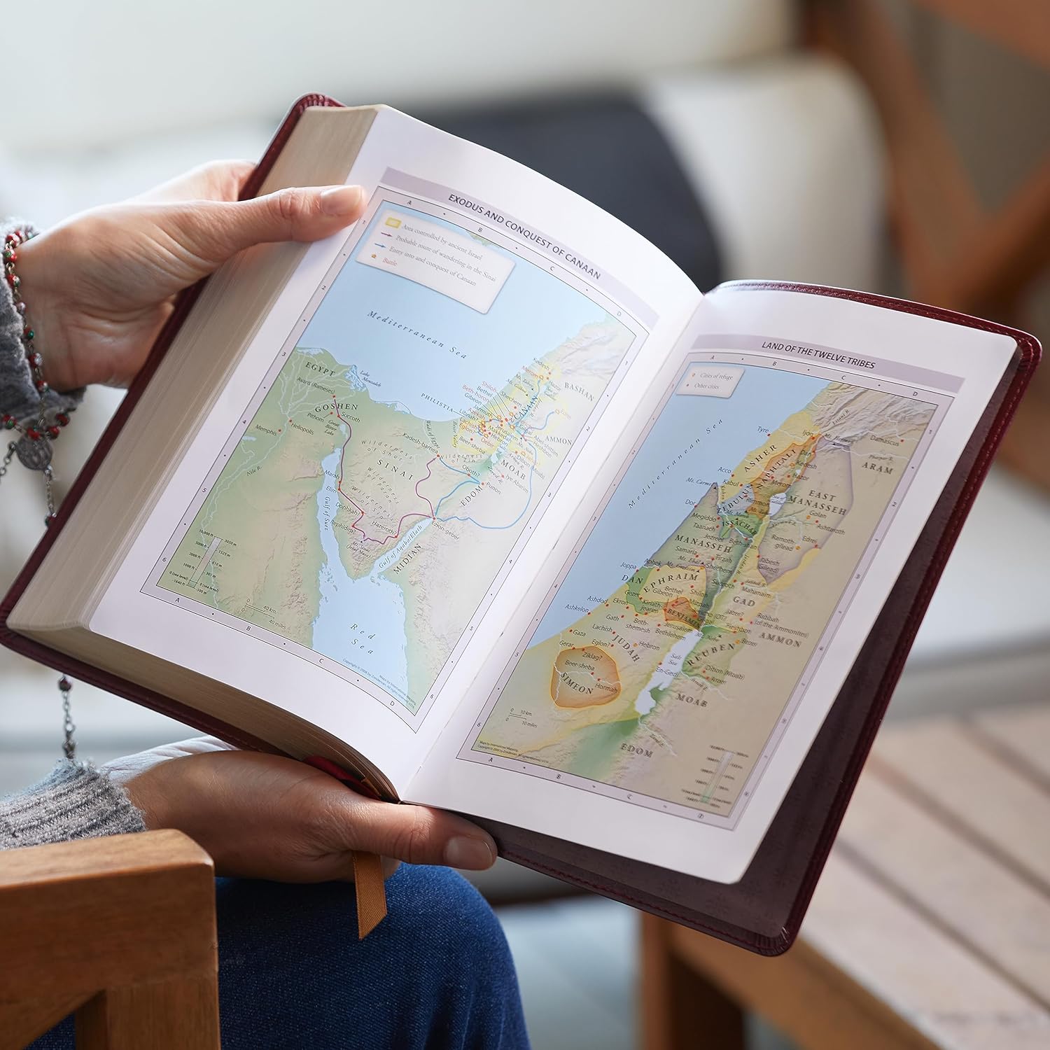 Open, burgundy leather Bible displaying colorful maps of Canaan and the Twelve Tribes in hands.