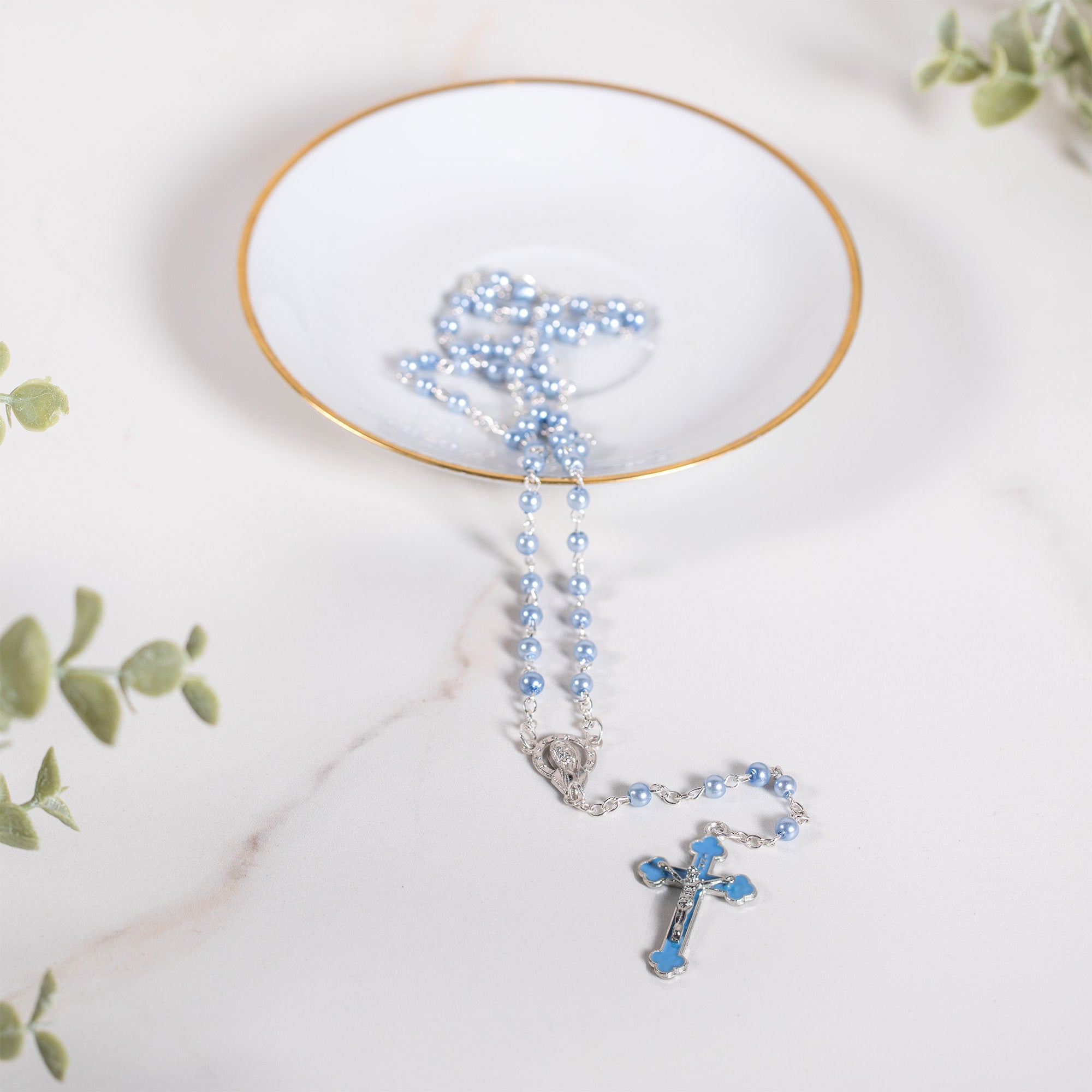 Light blue rosary with silver accents and cross pendant on a white and gold dish.