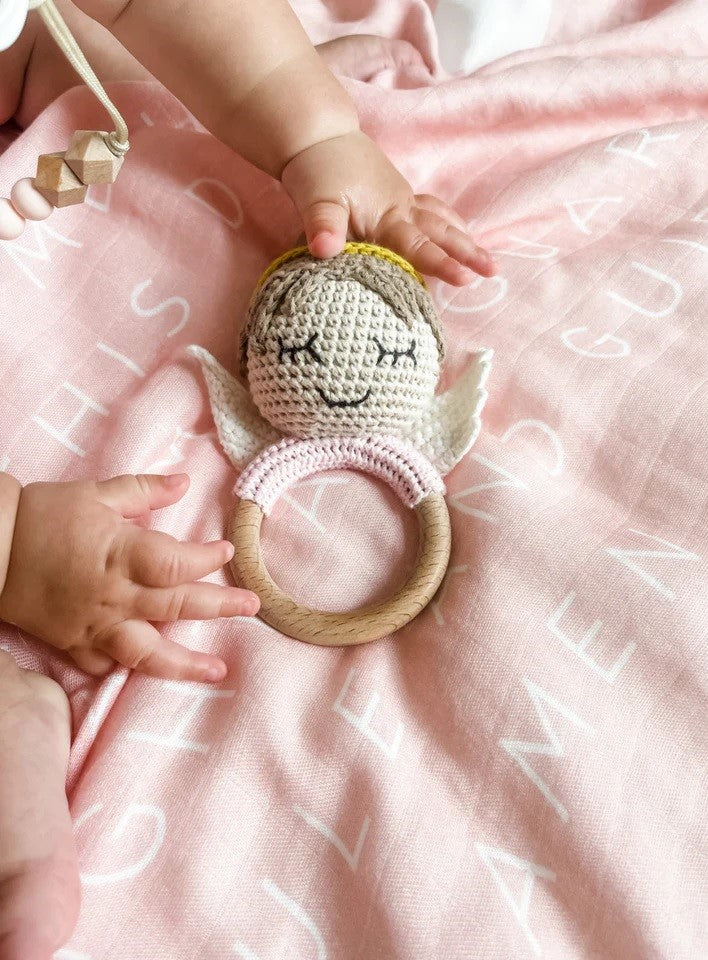Baby reaching for crochet angel teether with wooden ring and wings on a pink blanket.