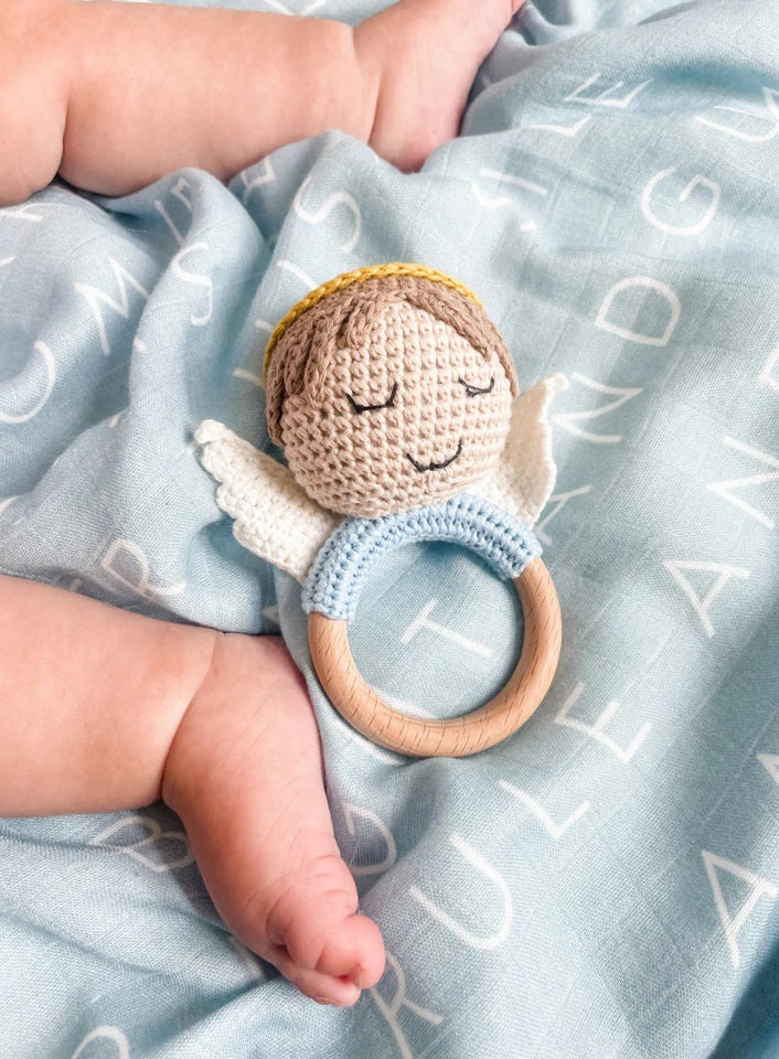 Crocheted angel baby rattle, with wooden ring, light blue accent, and white wings, on blue blanket with white letters.