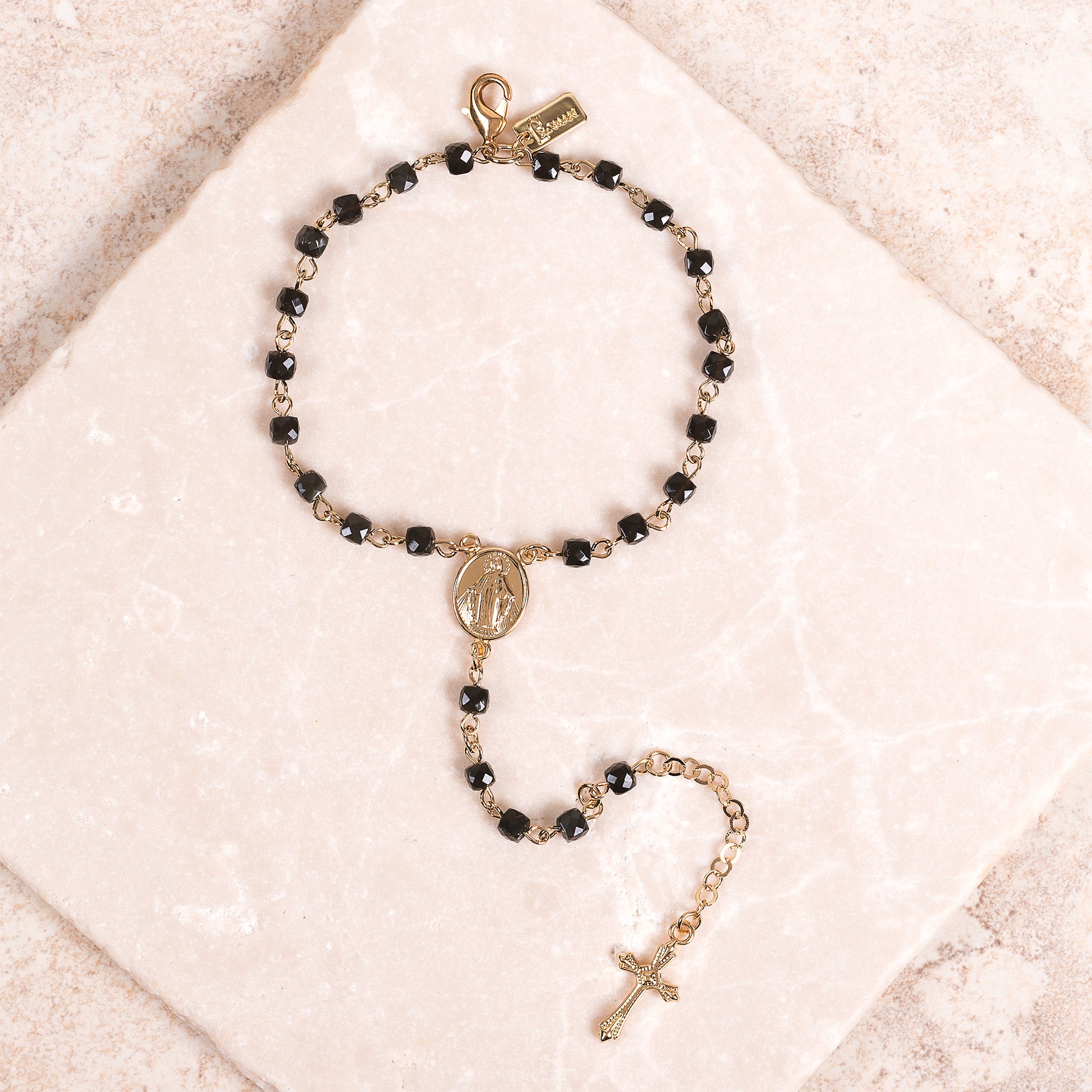 Rosary bracelet with black beads, gold chain, and crucifix charm, on a marble background.
