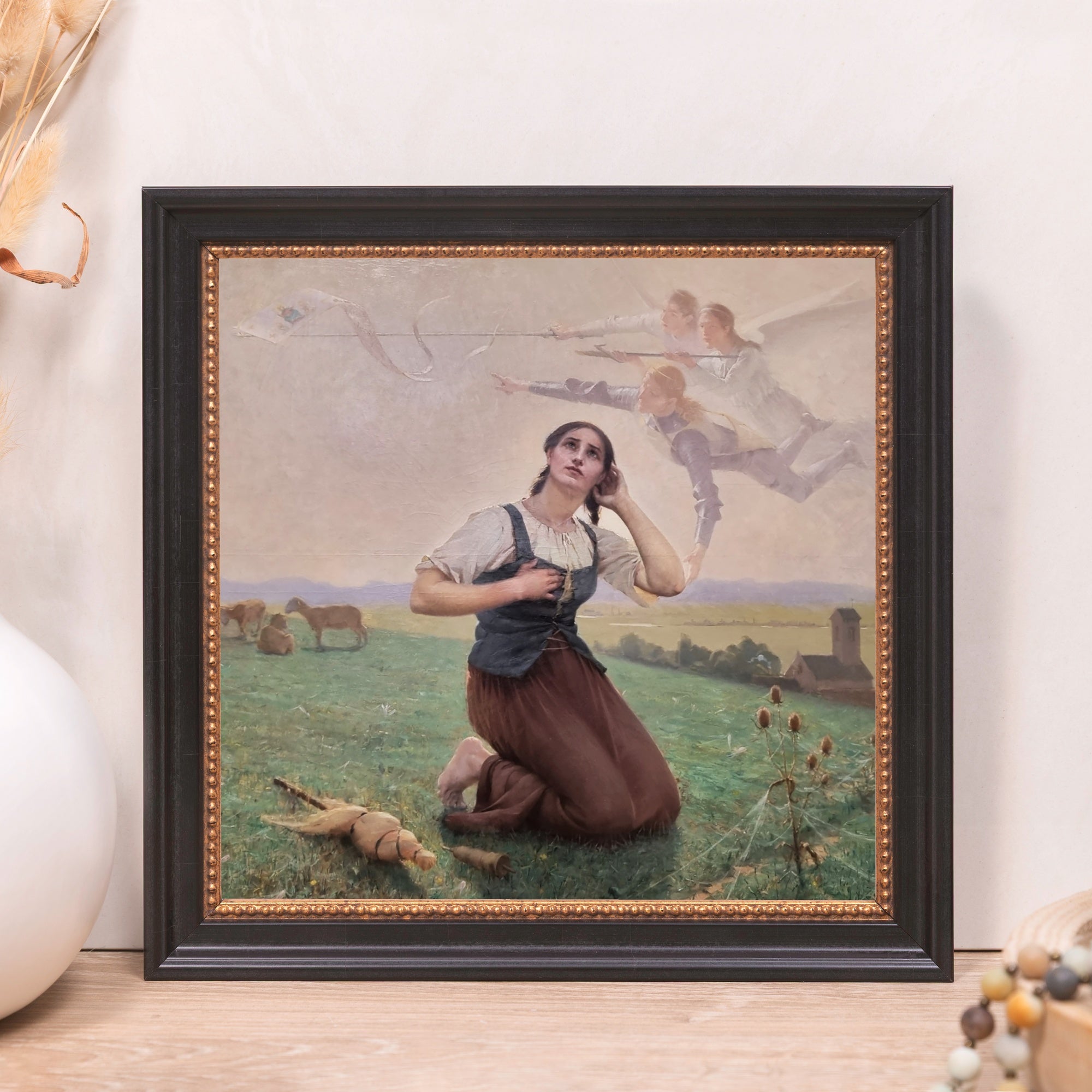 Framed painting depicts Joan of Arc in a field with angels overhead, in dark frame.