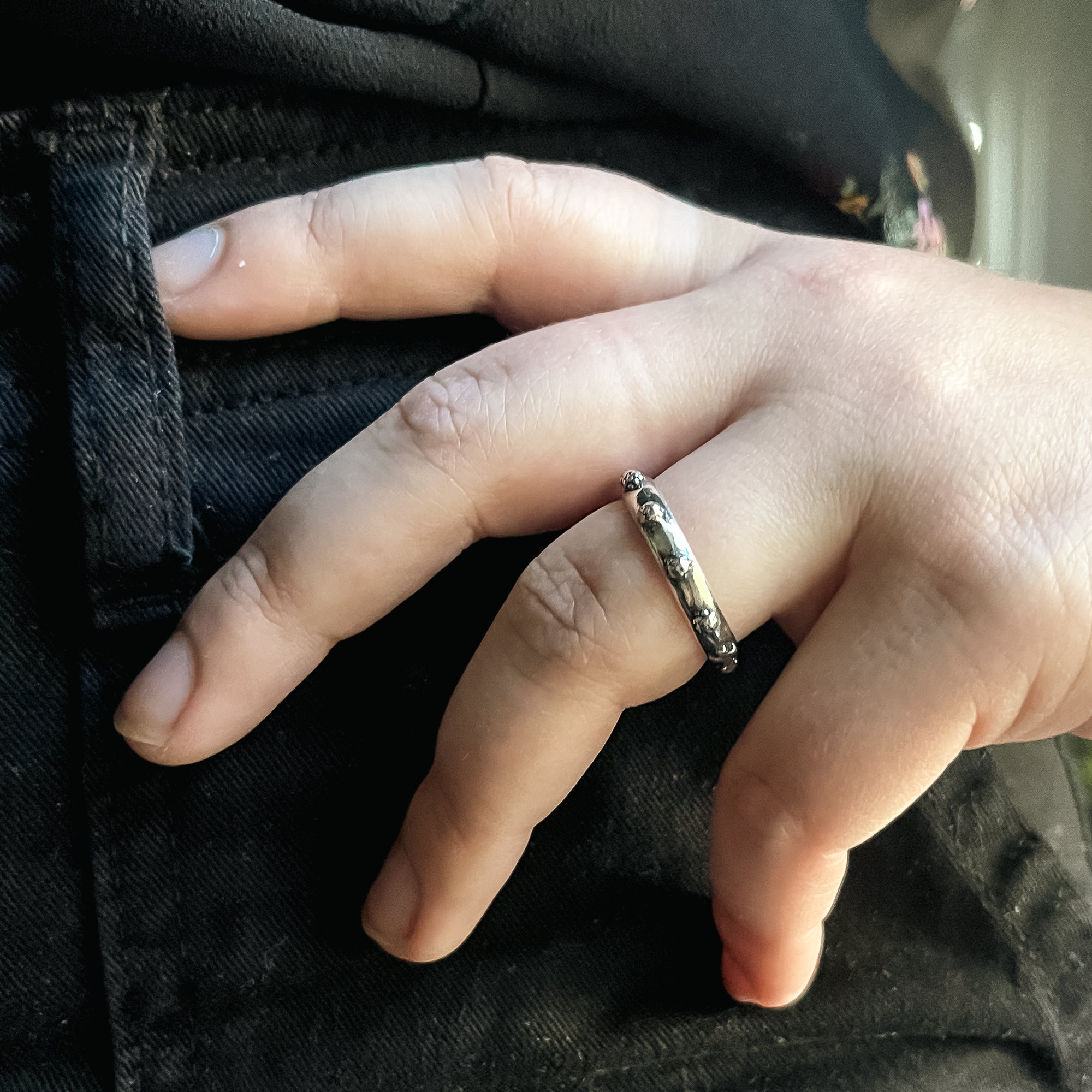 Silver ring on a hand over black fabric, has circular details all around it.