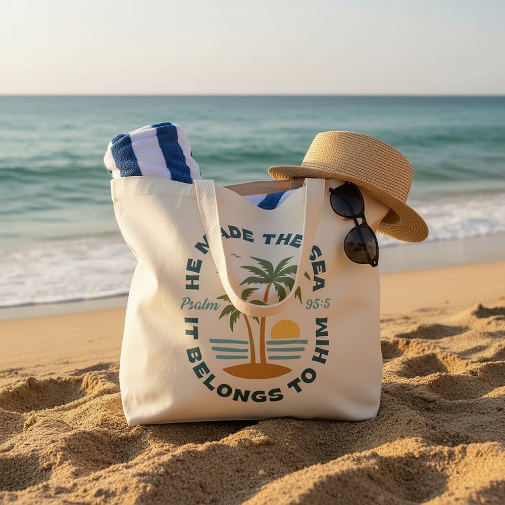 Canvas beach tote with palm tree design, lying on sand next to a straw hat and sunglasses.