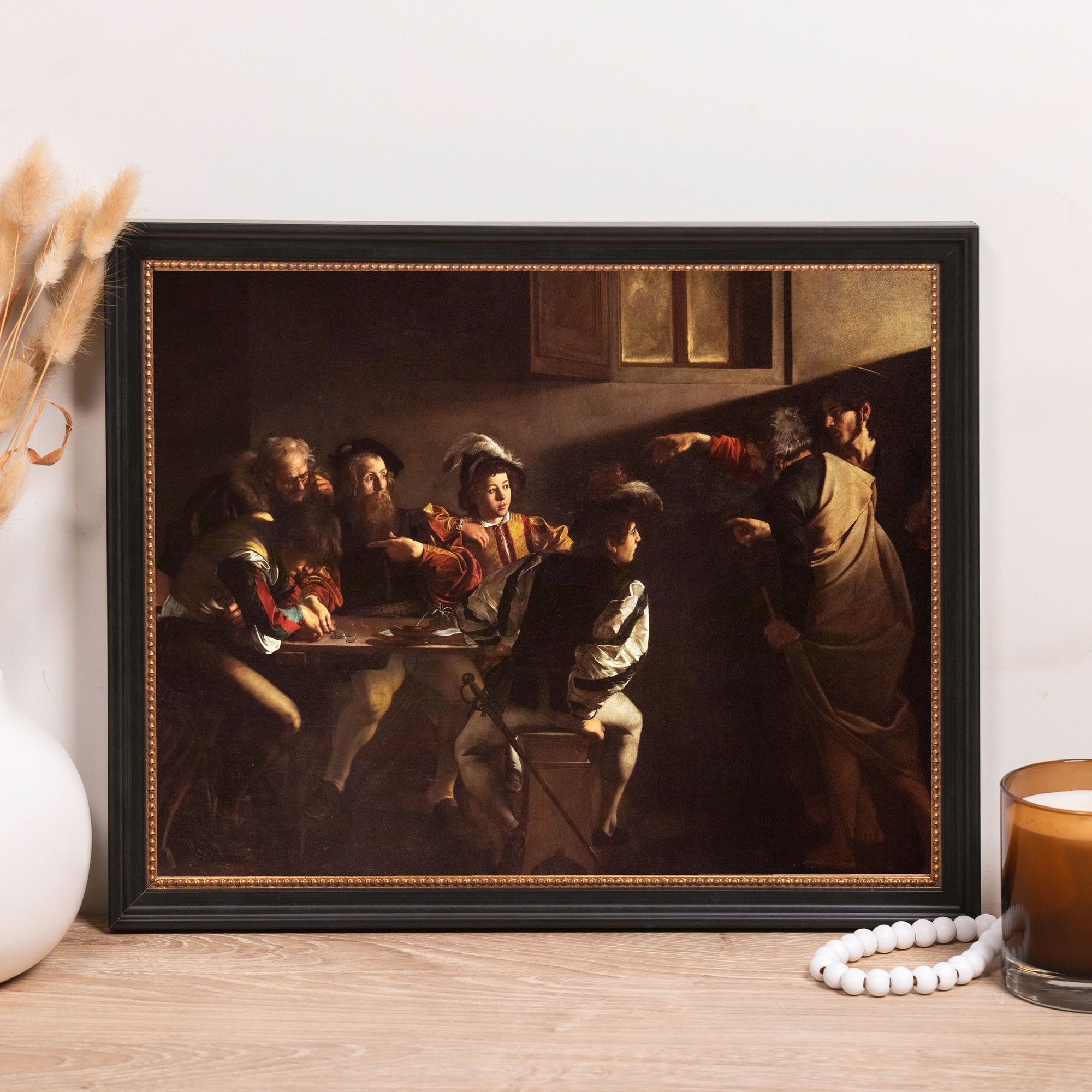 Framed print depicts "The Calling of St Matthew" by Caravaggio, in a dark palette, with a black frame.