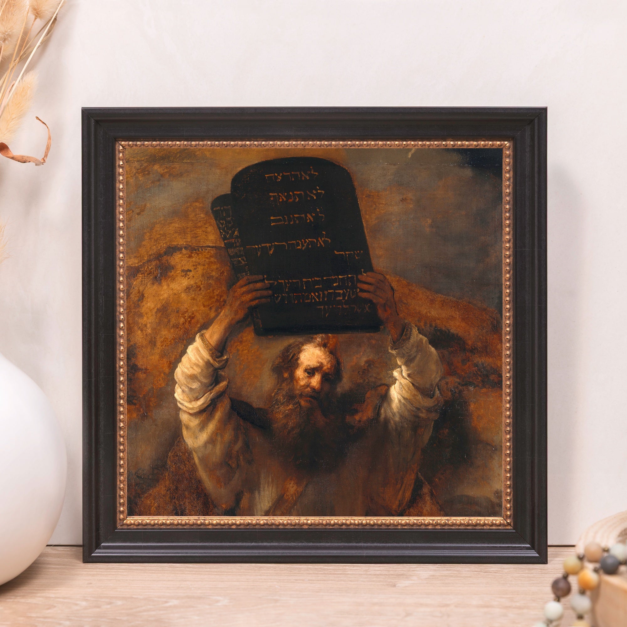 Framed painting shows Moses holding up the tablets of the law, written in Hebrew, in a black wooden frame with gold trim.