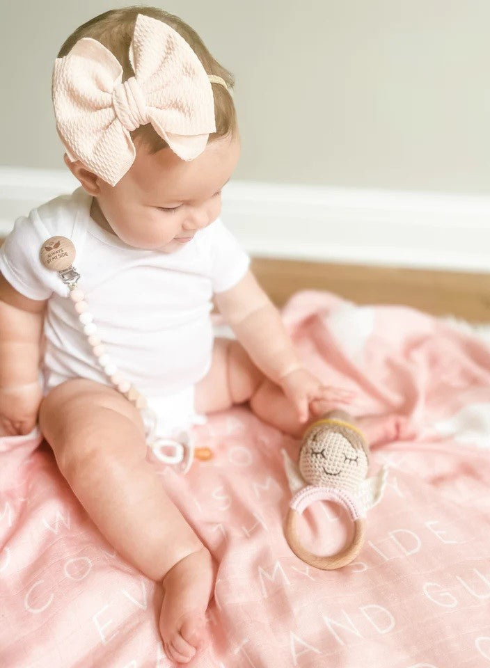 Baby with toy. Crocheted angel-shaped rattle with wood ring; baby wears a large pink bow and "Always By My Side" pacifier clip.