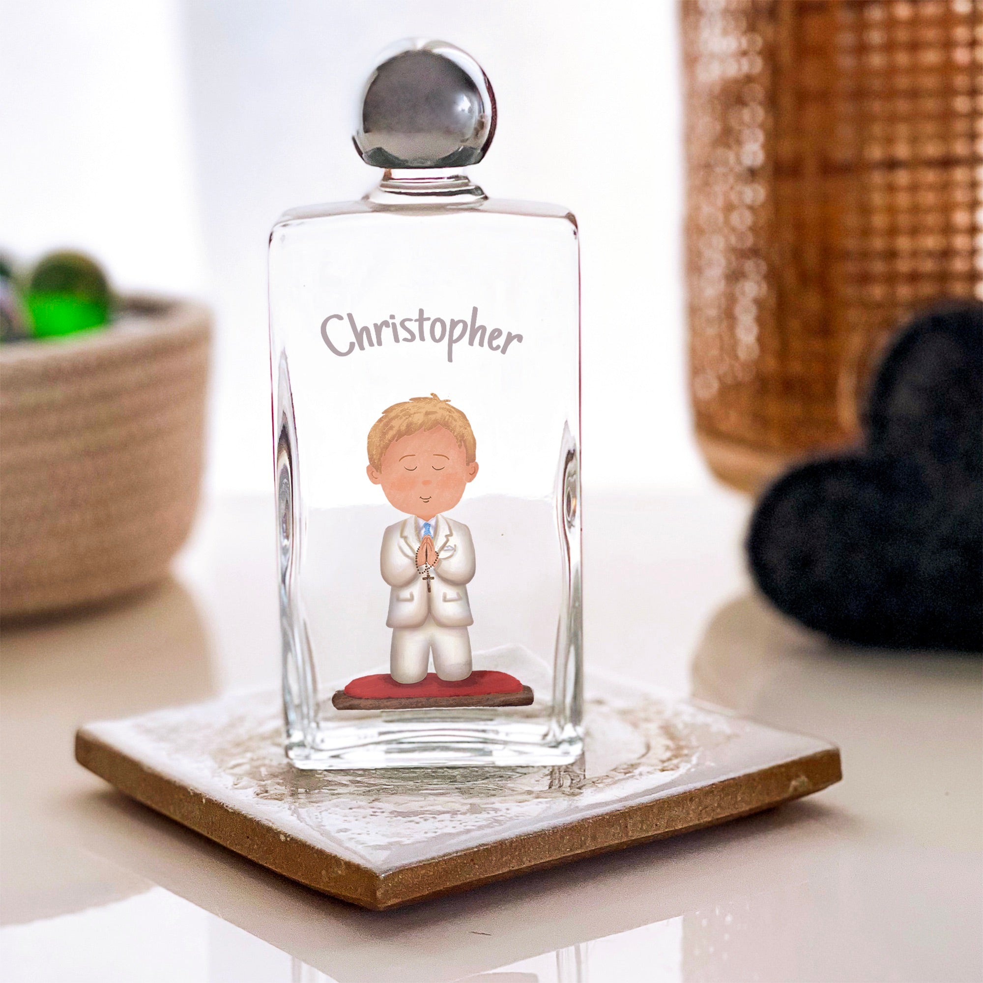 Personalized, clear glass holy water bottle with gray text and communion boy design; placed on a patterned coaster.