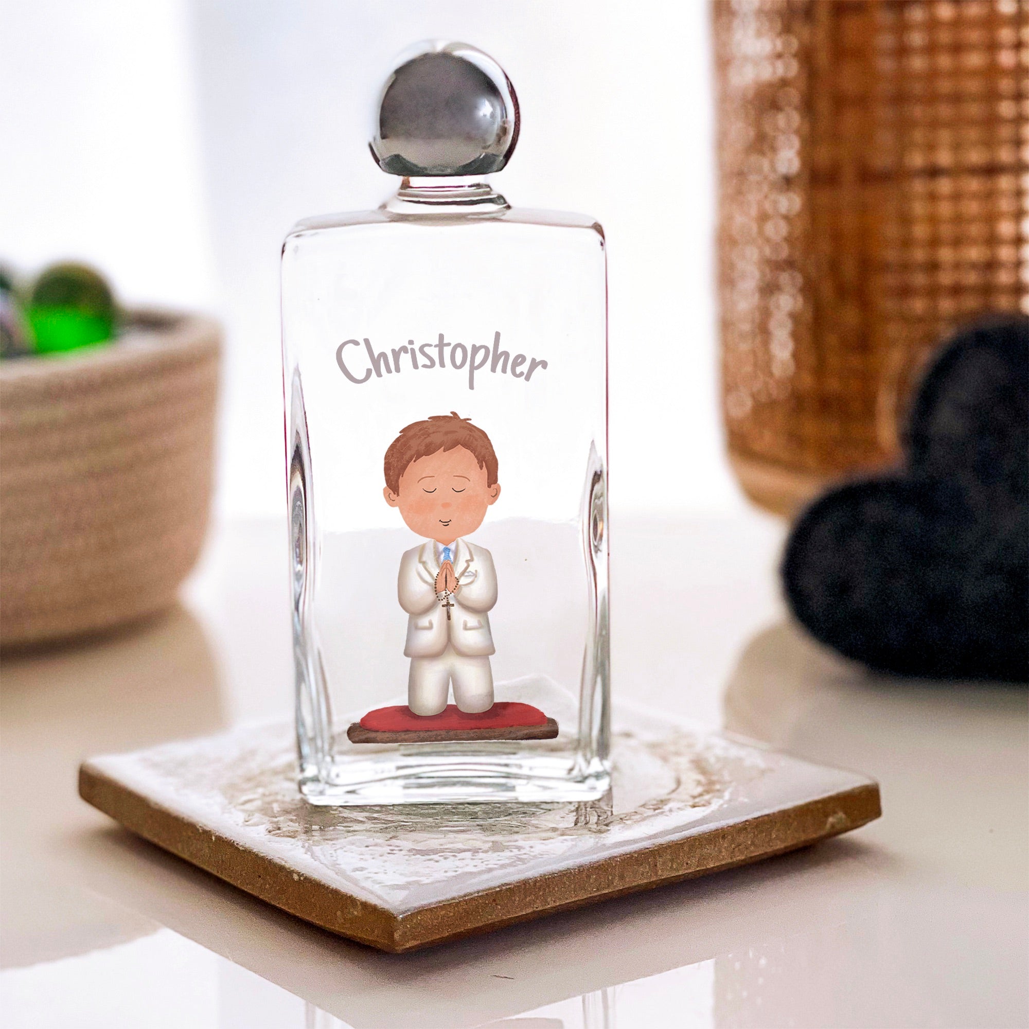 Personalized holy water bottle with a boy illustration, clear glass and silver cap, on a tile coaster.