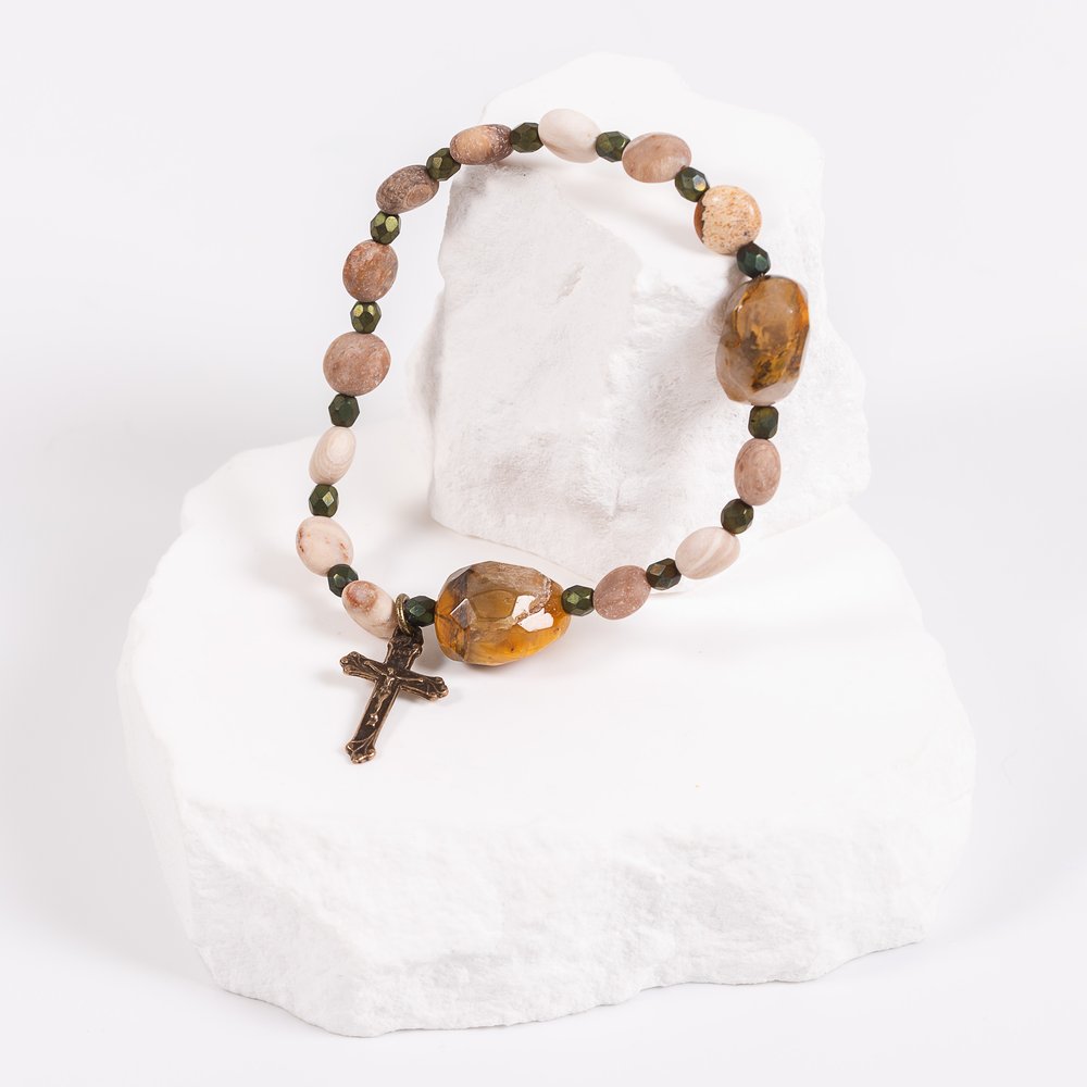 Beaded bracelet with a cross charm, featuring earthy-toned stone beads and green accents, on white display stand.
