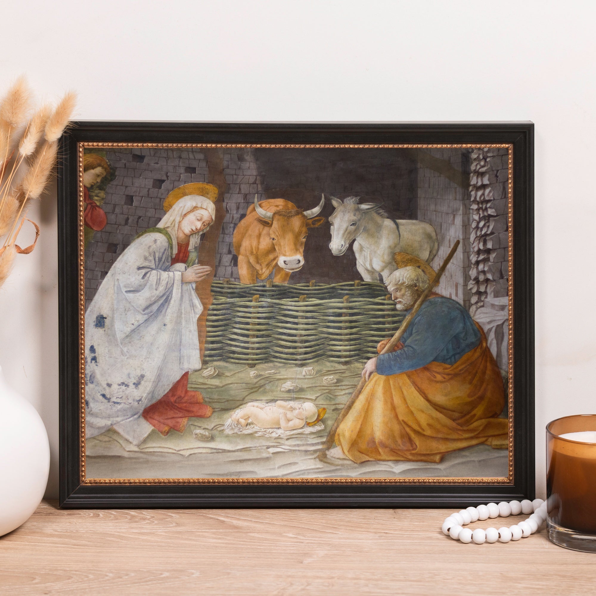 Framed print of the Nativity scene in a vintage style, displayed on a wood shelf with a white vase and candle.