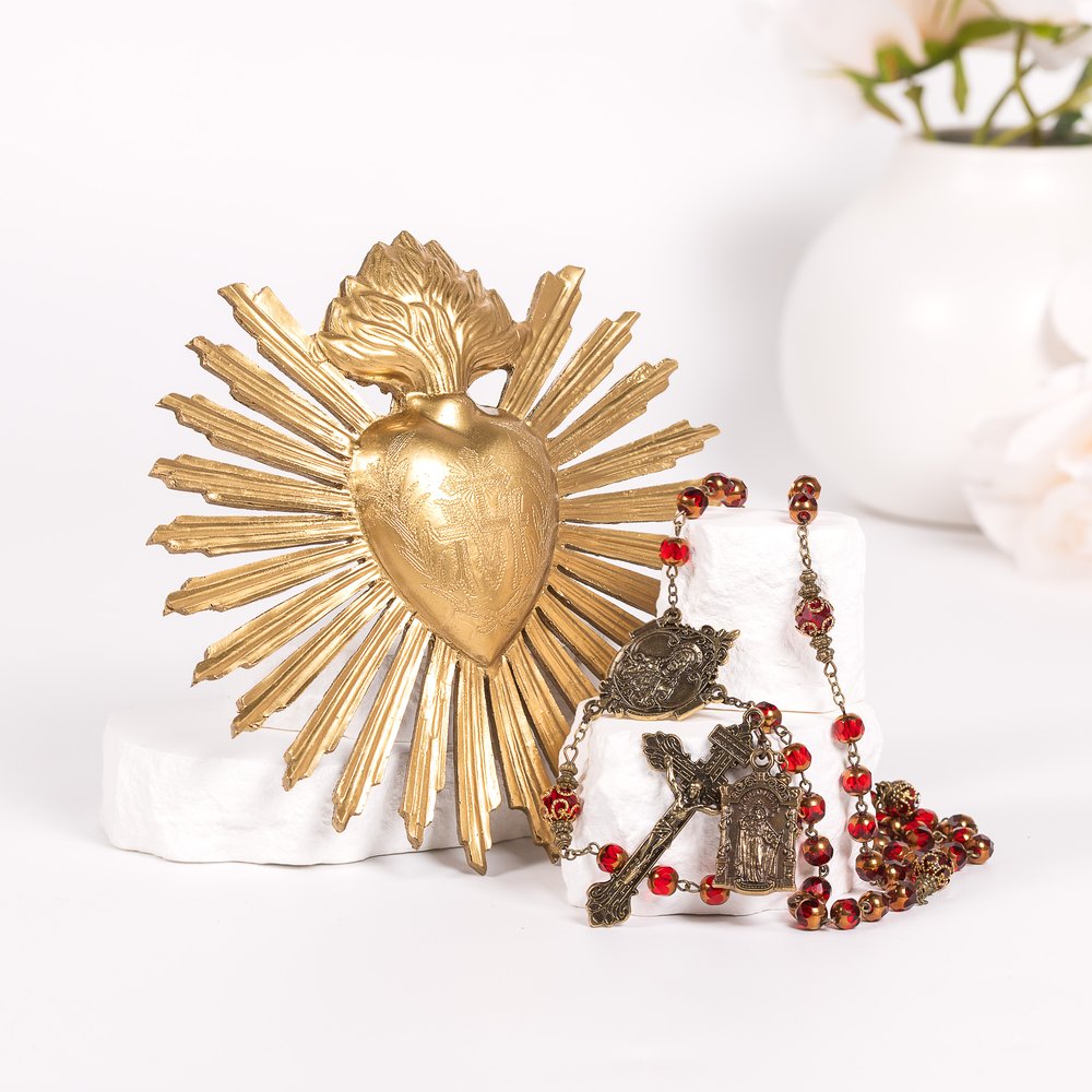 Golden, heart-shaped Sacred Heart wall decor with rays and a red-beaded rosary with a metal cross on a white surface.