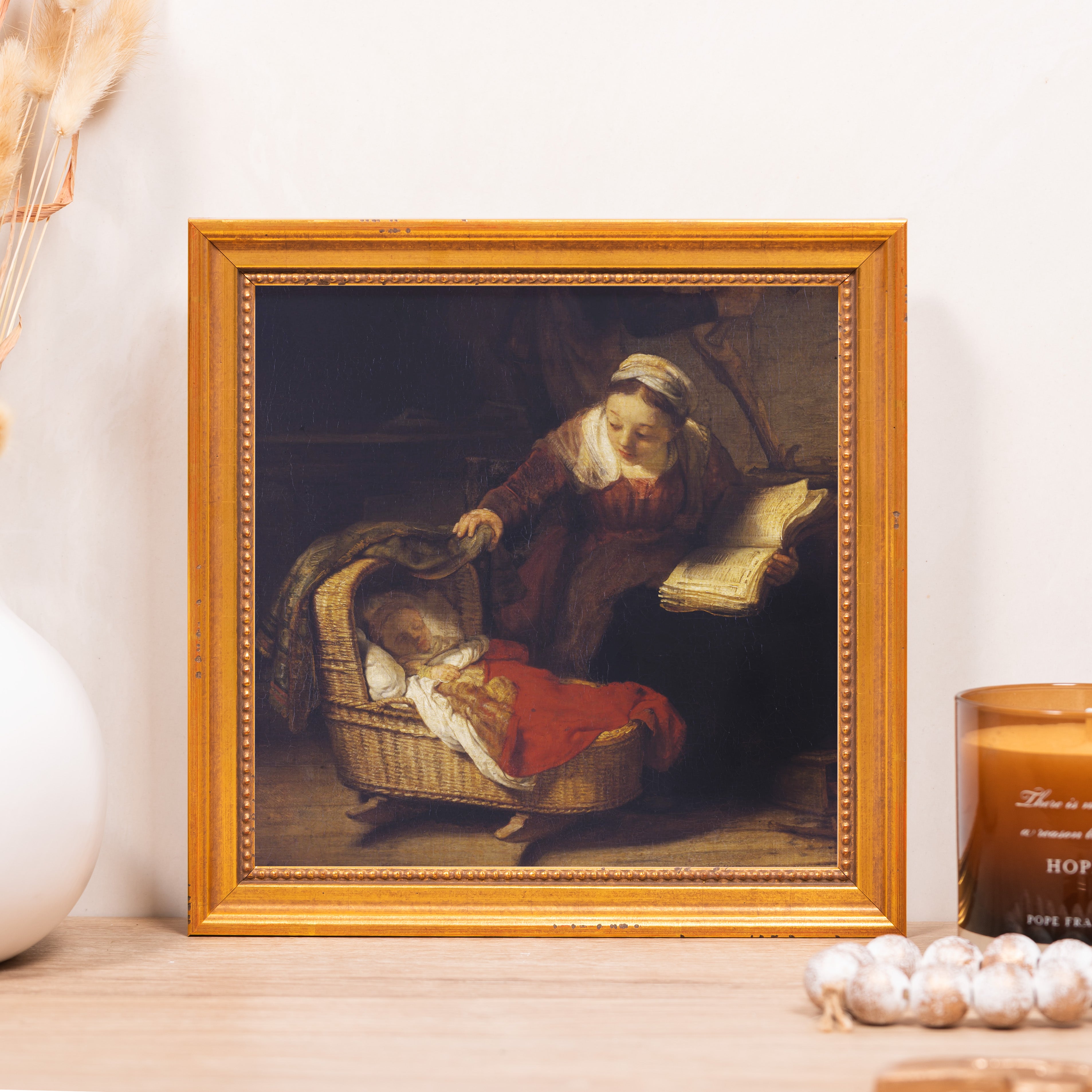 Framed oil painting depicting a woman reading beside a woven baby cradle, in a gold frame.
