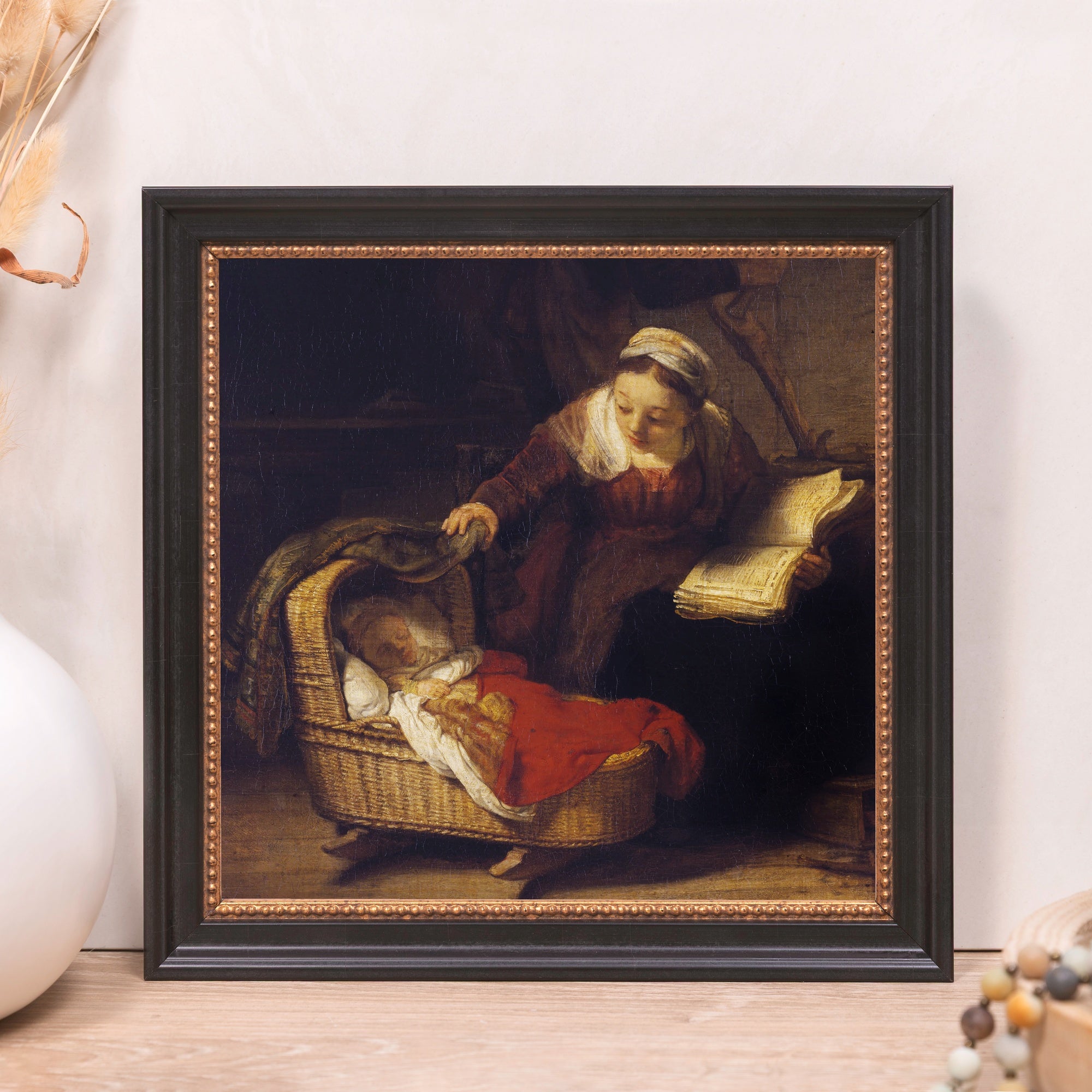 Framed reproduction of Rembrandt's "Mother Reading Beside a Cradle," in a dark wood frame with gold-tone inner trim.