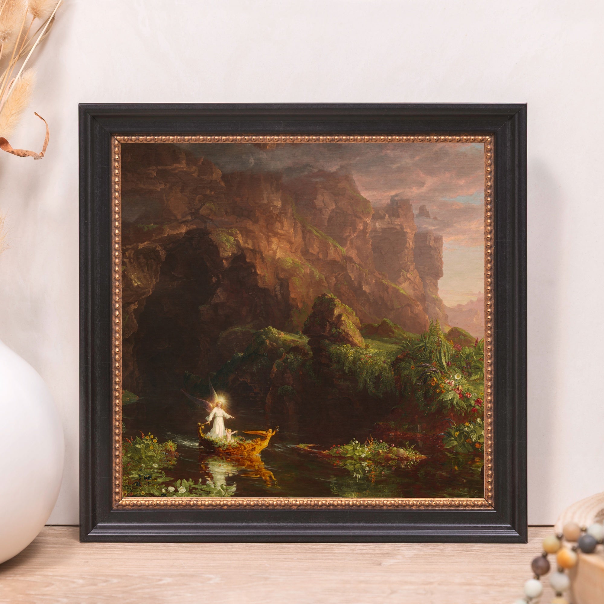 Framed landscape painting of figures in a boat on a river with trees and cliffs in a black and gold frame.