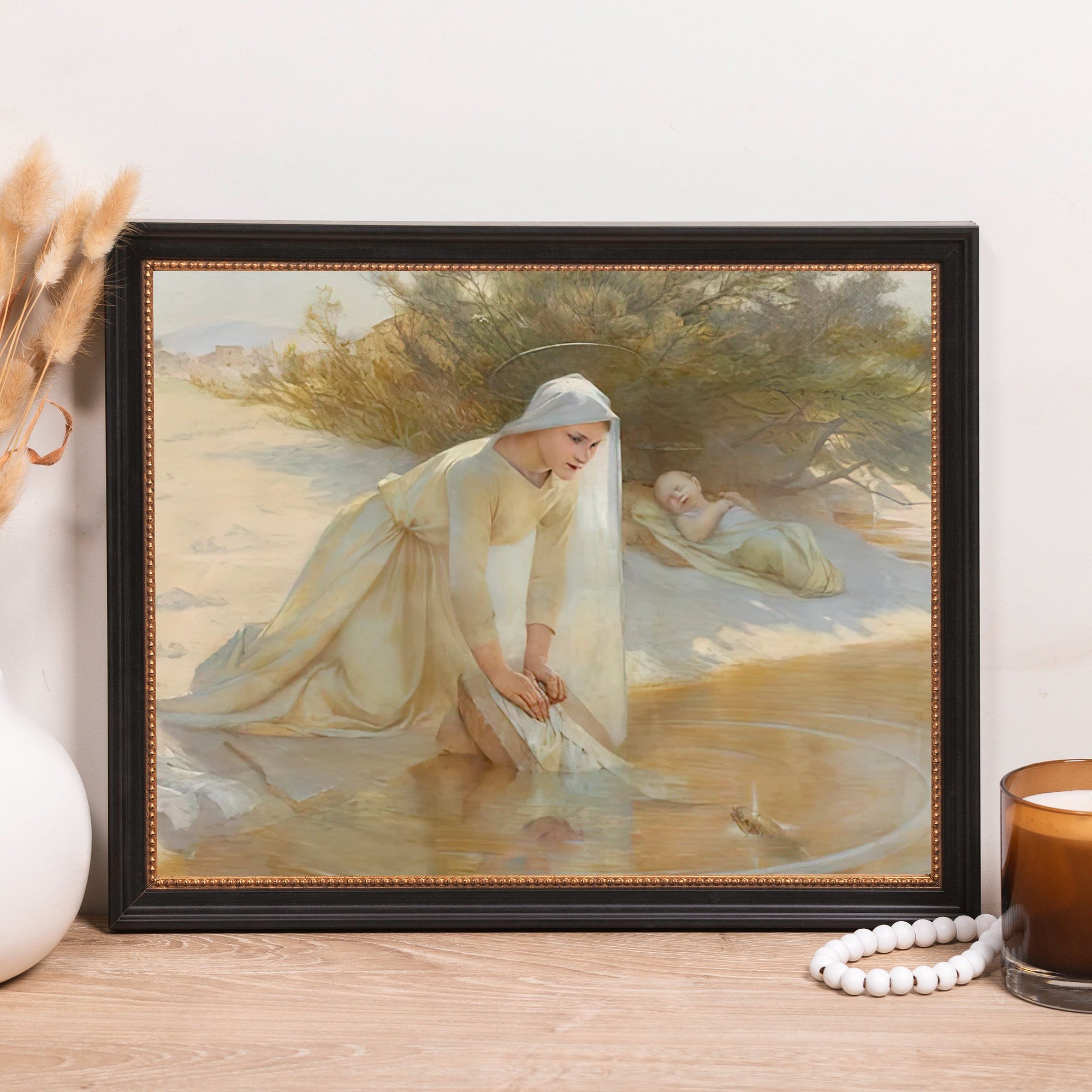 Framed oil painting of Mary washing Jesus in a river, light colors, in a dark frame.