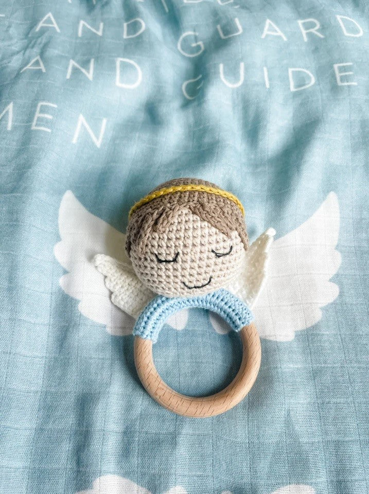 Crocheted angel baby rattle with wooden ring, light blue trim, and white wings on a blue blanket.