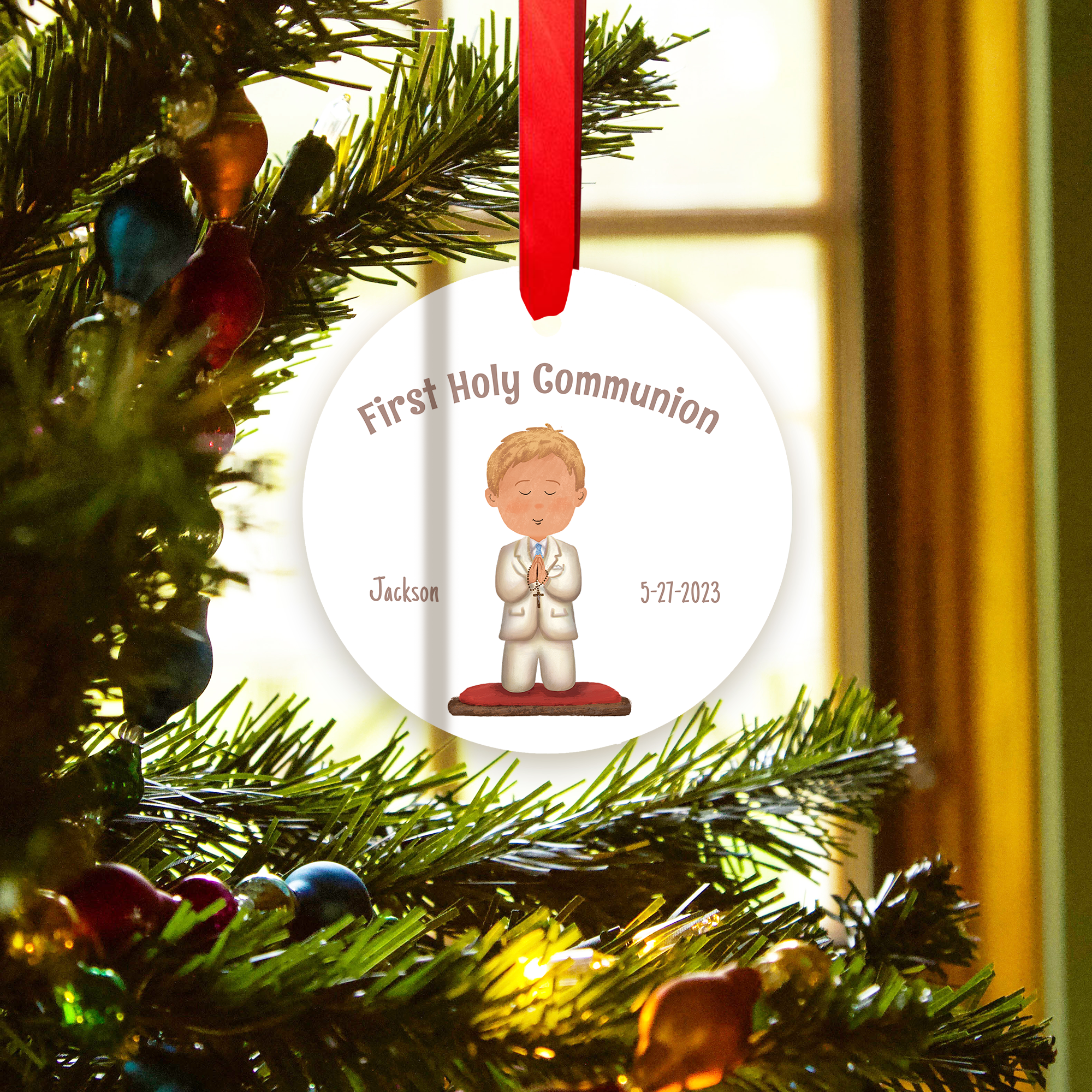 White, round ornament for First Holy Communion, with red ribbon, hangs on a tree.