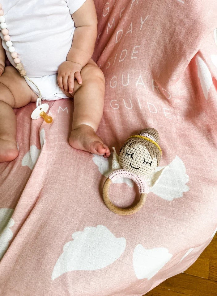 Crochet Angel baby rattle made with light brown yarn, wood ring, on a blush pink blanket with white clouds and text.