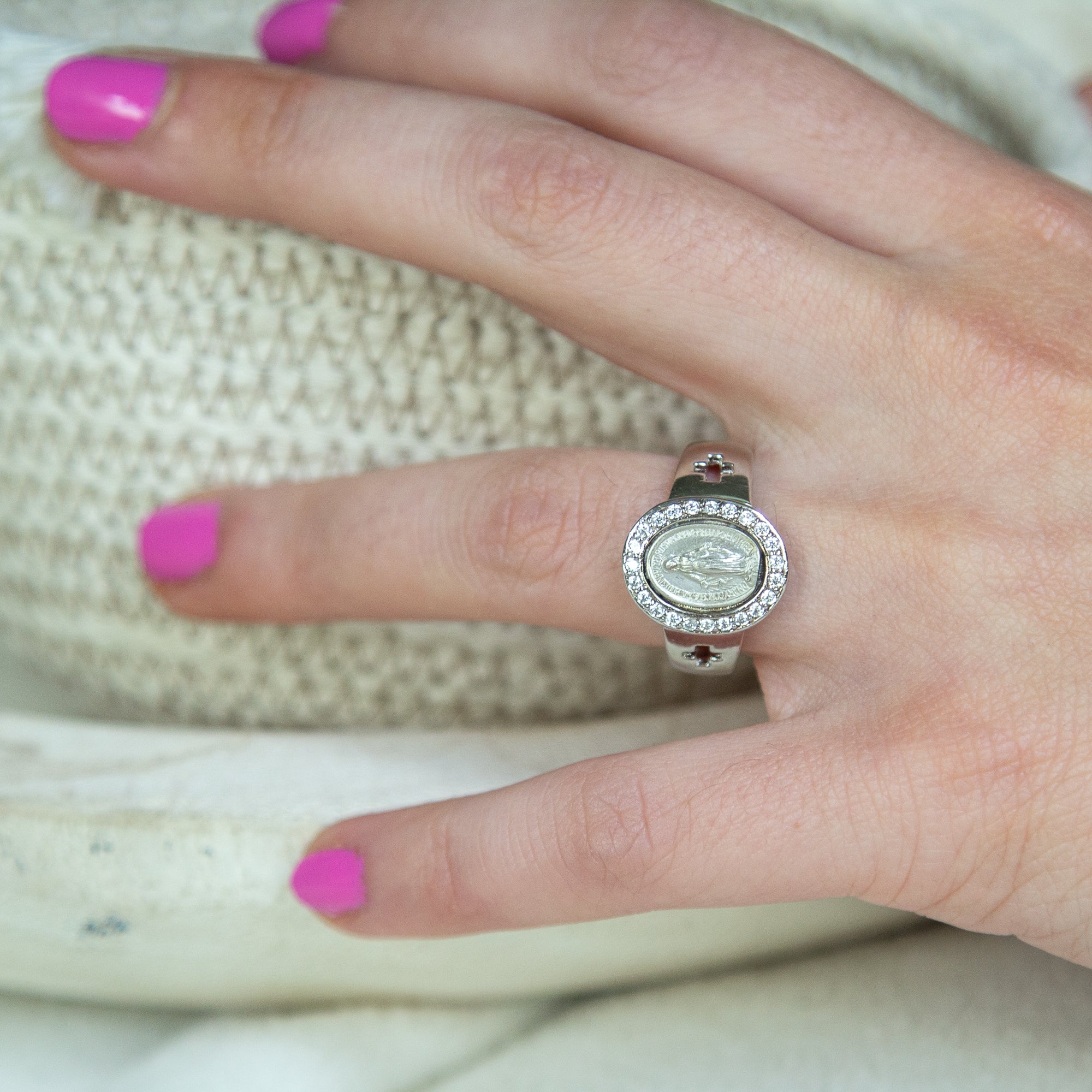 Silver ring with an oval Virgin Mary centerpiece surrounded by clear rhinestones on a hand with hot pink nail polish.