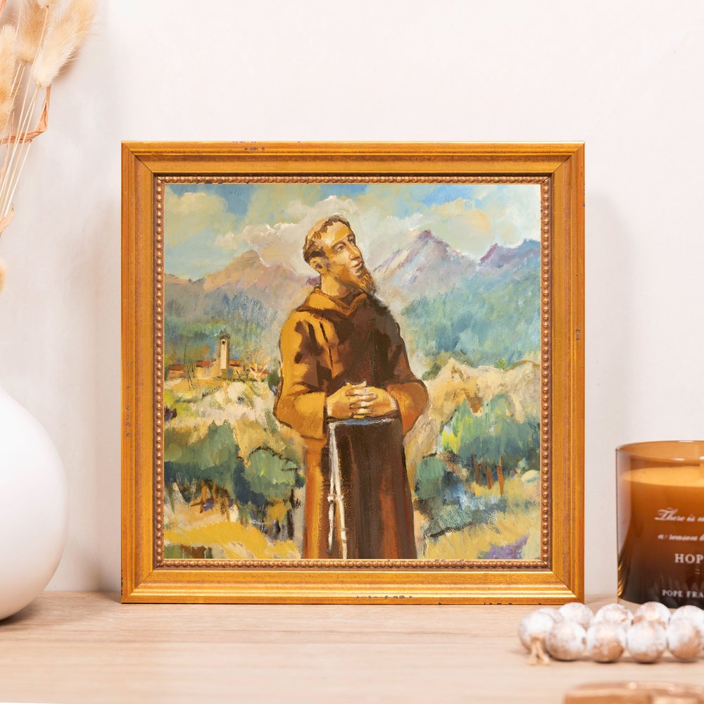 Framed religious painting depicting Saint Francis with a brown robe, painted landscape background, and gold frame.