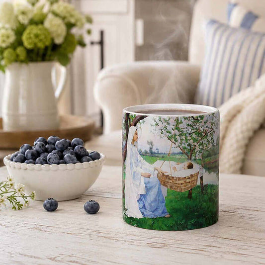A ceramic mug with a printed illustration of a woman in a blue dress beside a basket holding a baby, placed on a wooden table in a softly styled living room setting with blueberries and flowers in the background.