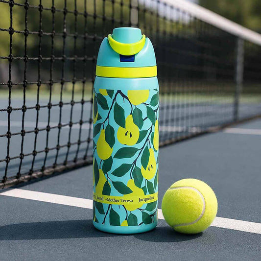 A reusable water bottle with a turquoise and yellow lemon pattern stands on a tennis court next to a tennis ball. The bottle has a matching turquoise and yellow flip-top lid.