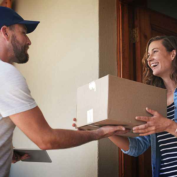 Man in a blue hat handing a smiling woman standing in a doorway, a cardboard box.